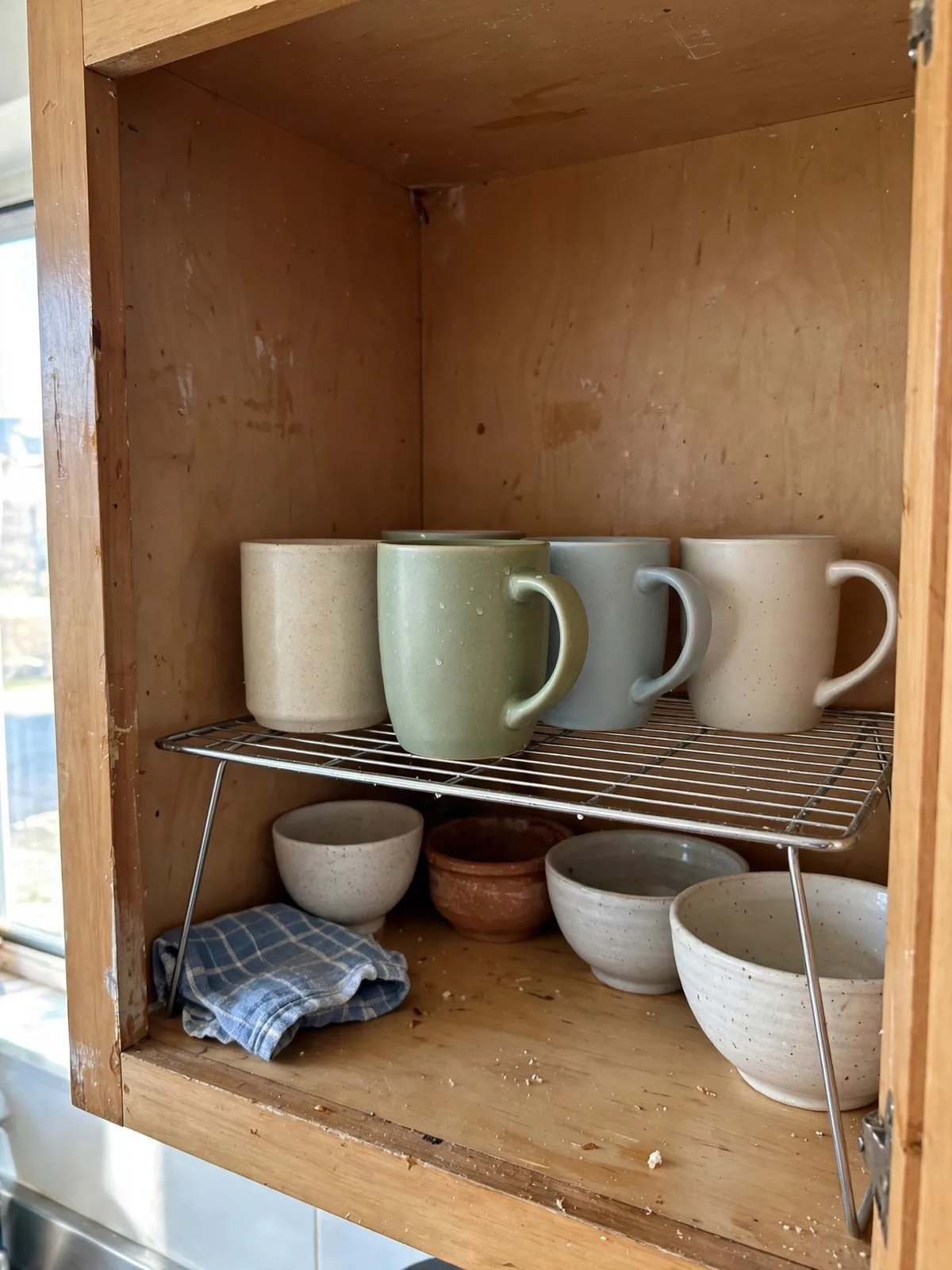 Shelf risers inside a small apartment kitchen cabinet splitting one shelf into two levels, with mugs and bowls arranged neatly