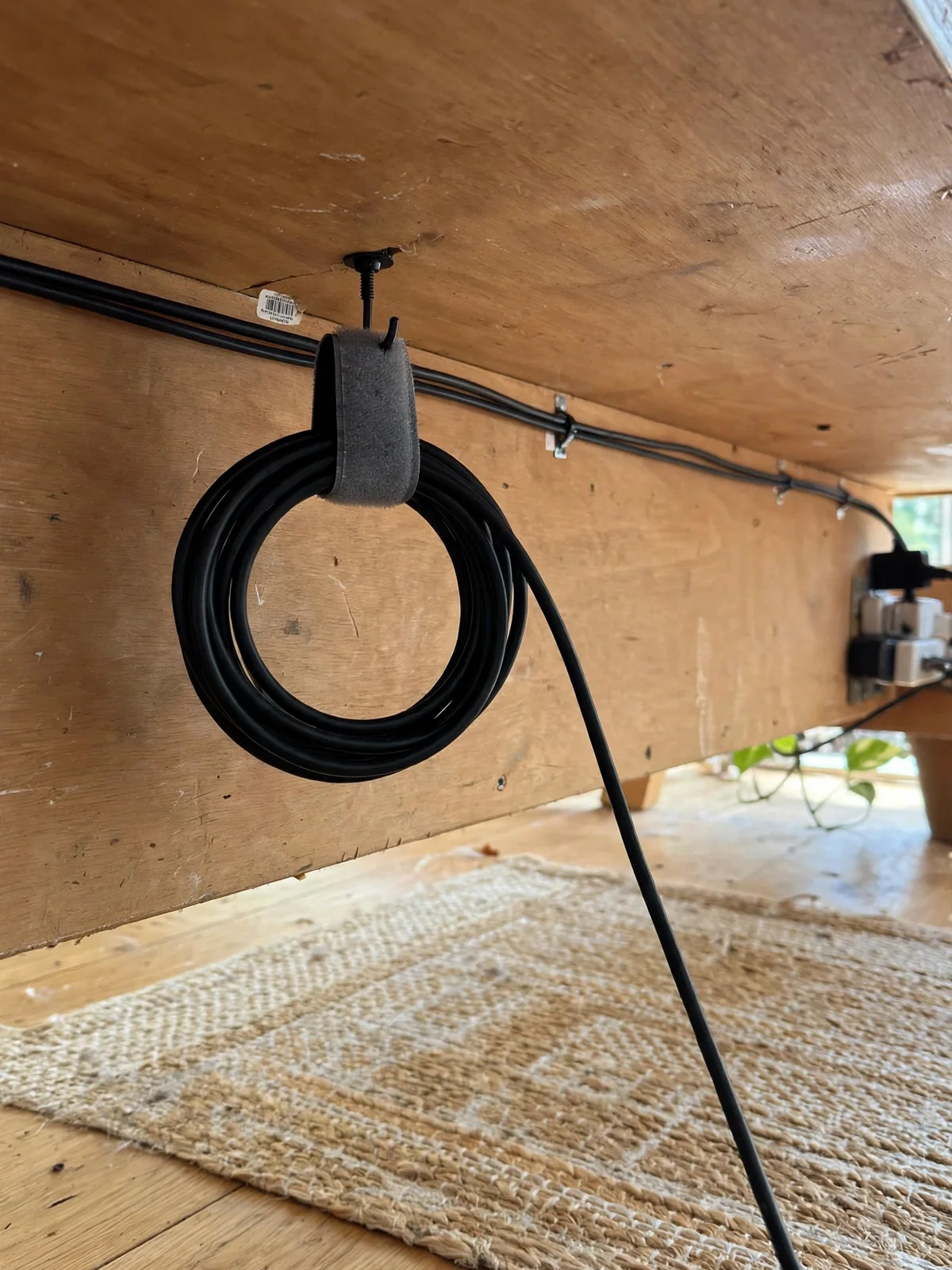 Excess black cable coiled into a neat loop and hung from a hook under a wooden desk with a grey Velcro strap, floor below completely clear