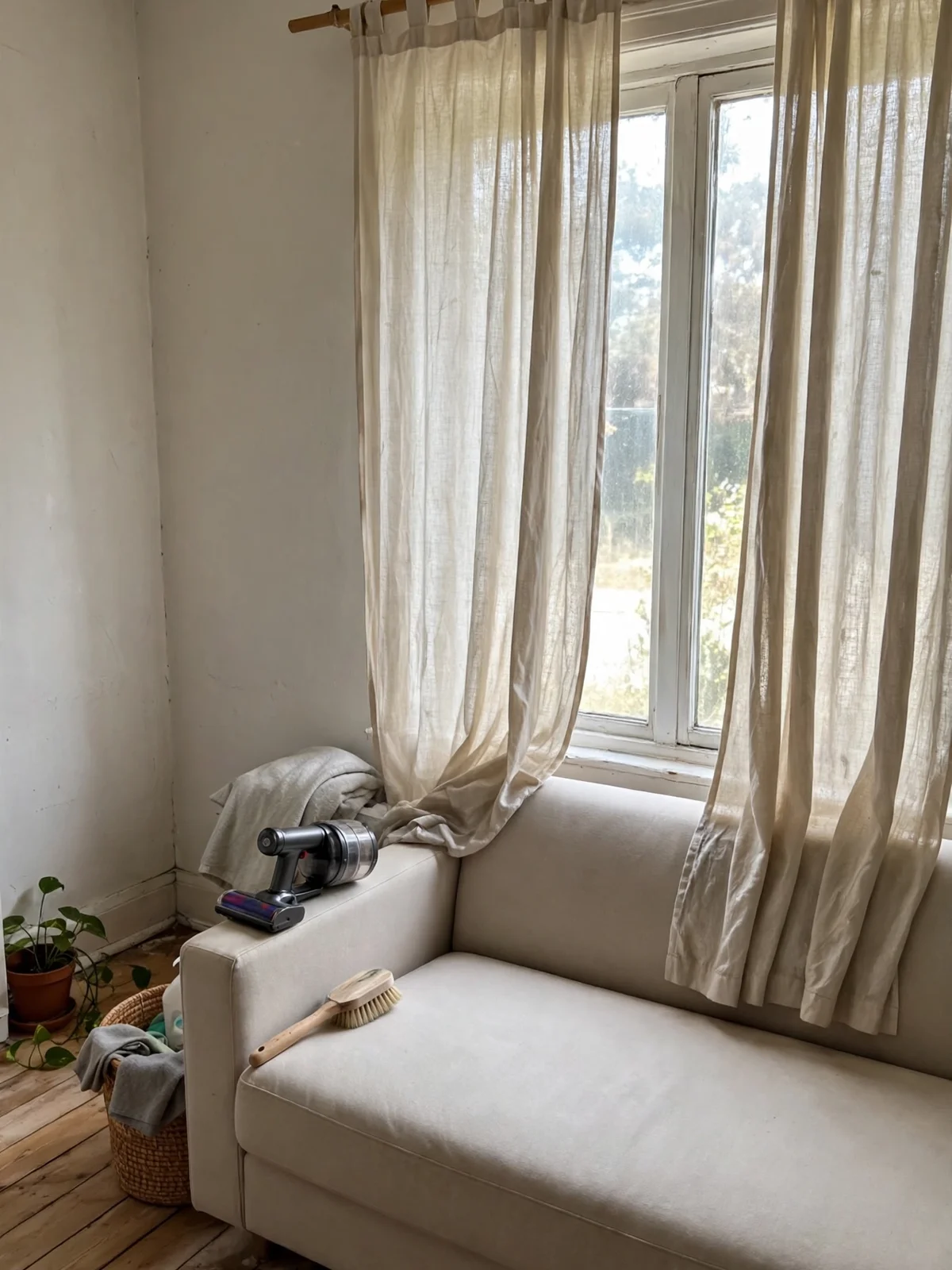 Freshly washed linen curtains hanging by a bright window above a vacuumed fabric sofa with an upholstery brush and handheld vacuum on the arm