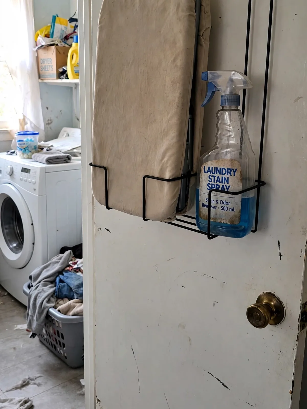 Ironing board stored upright in an over the door holder on a laundry room door with a spray bottle beside a washing machine nearby