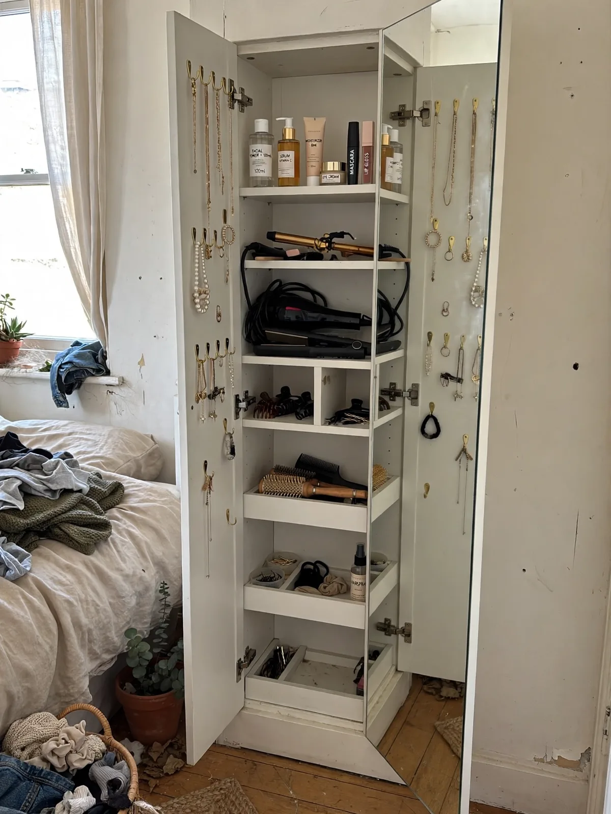 Full length mirror on a bedroom wall swung open to reveal a shallow cabinet inside with jewelry hooks makeup bottles and hair tools stored