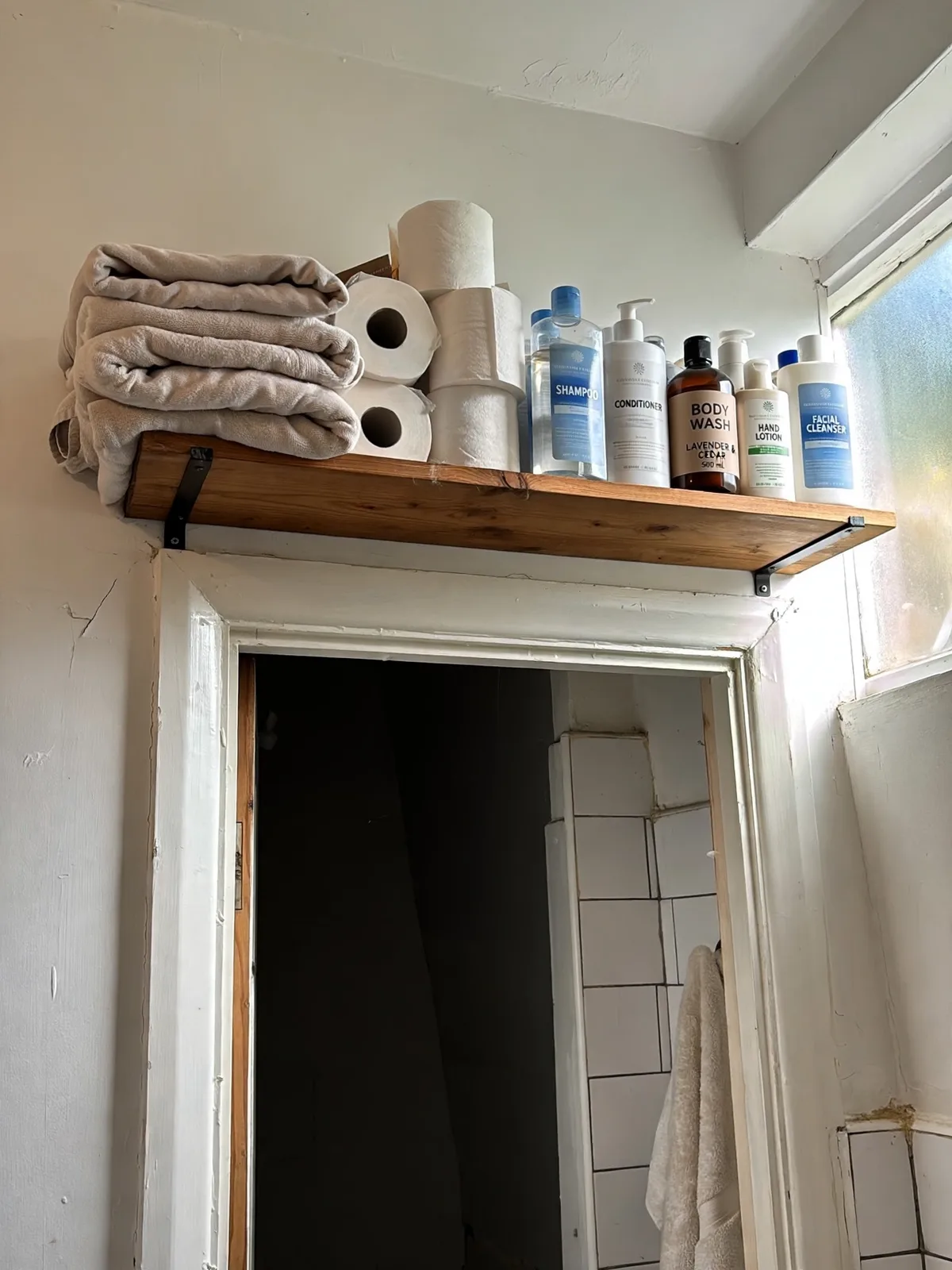 Simple wooden shelf mounted above a doorway inside a small bathroom holding folded extra towels toilet paper rolls and stock up toiletry bottles neatly