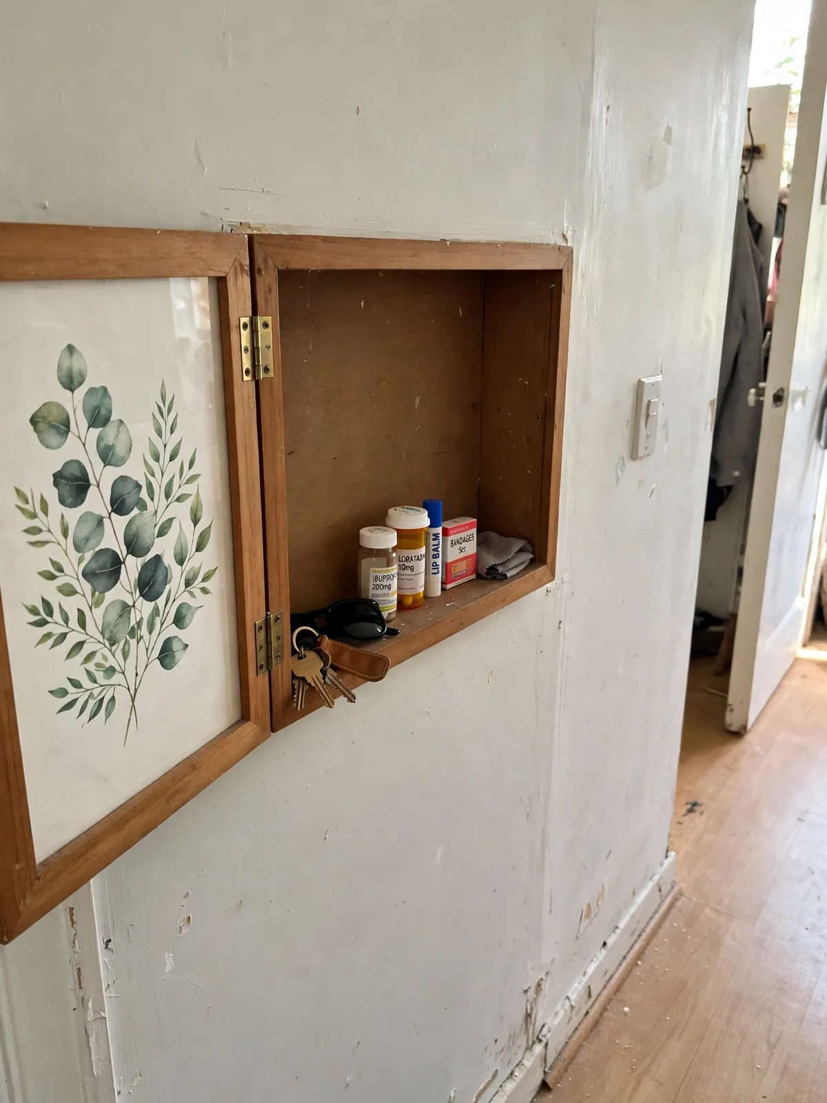 Framed artwork on a hallway wall opened outward revealing a shallow hidden cabinet behind it holding keys sunglasses medicines and small everyday items inside