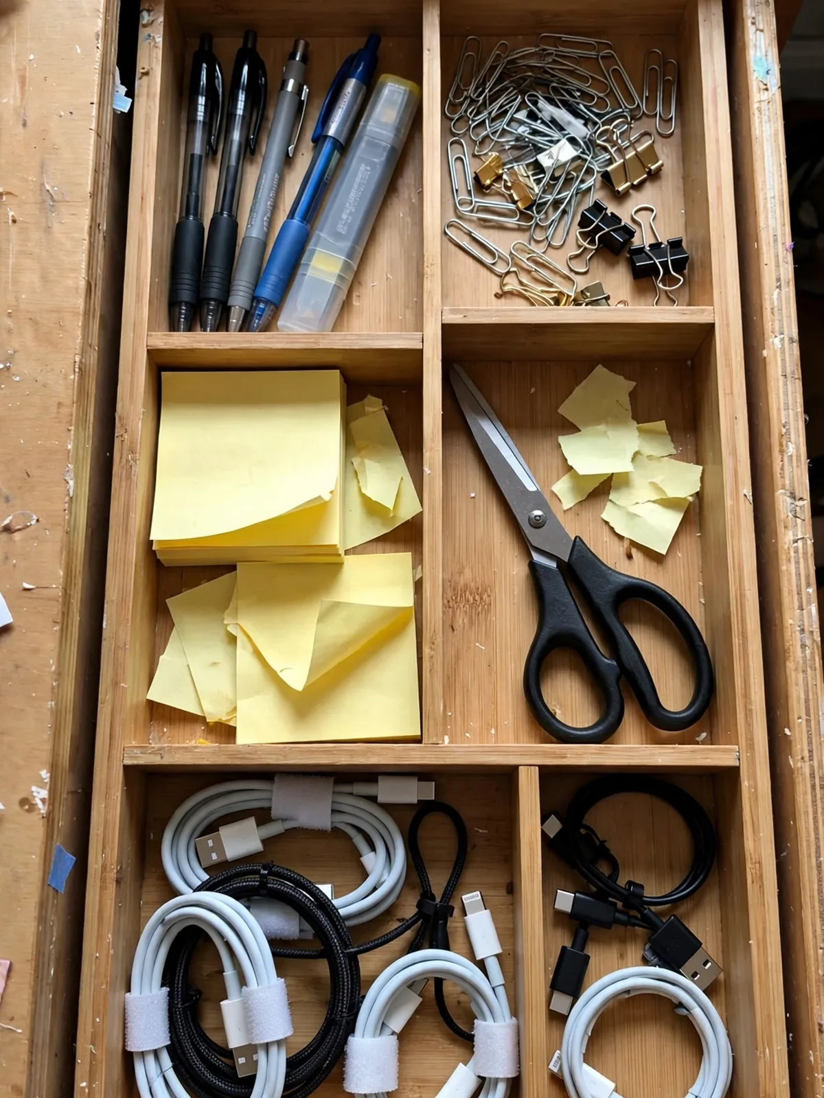 Open shallow desk drawer from above with a bamboo divider tray organizing pens, paper clips, sticky notes, scissors and cables into neat sections