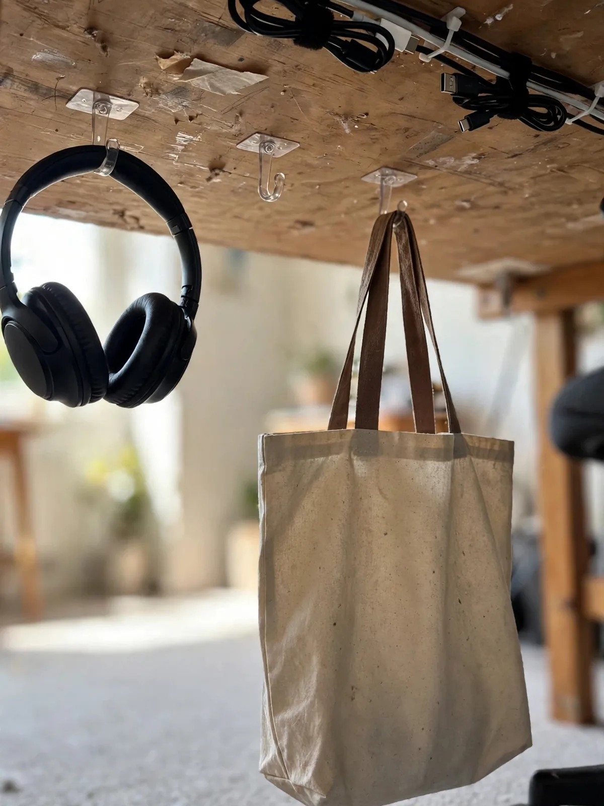 View under a small desk showing adhesive hooks holding headphones and a tote bag off the floor with tidy cables above them