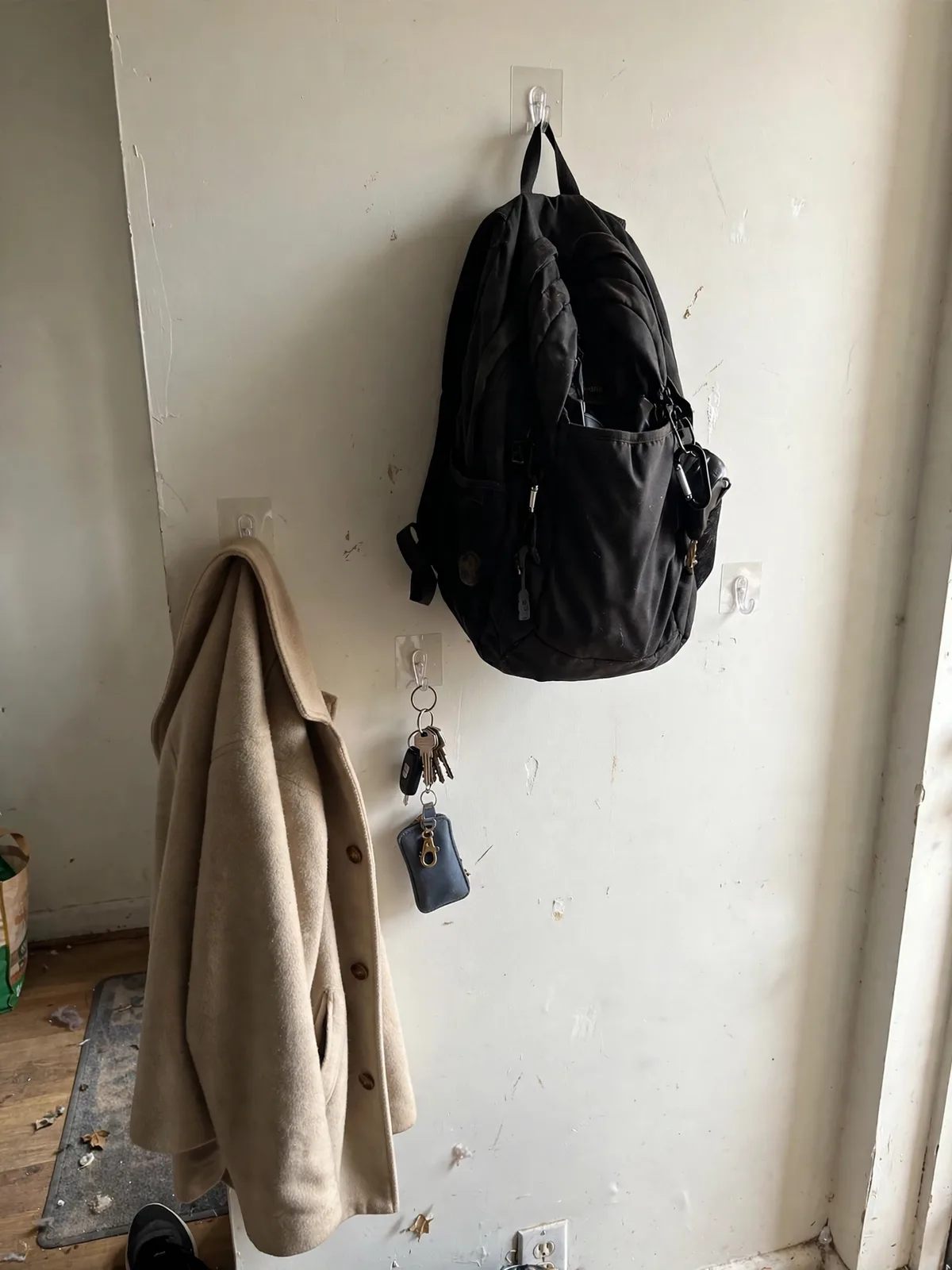 Entryway wall in a small rental apartment with clear adhesive hooks holding a coat, backpack and keys, stuck on painted wall with no visible holes or damage