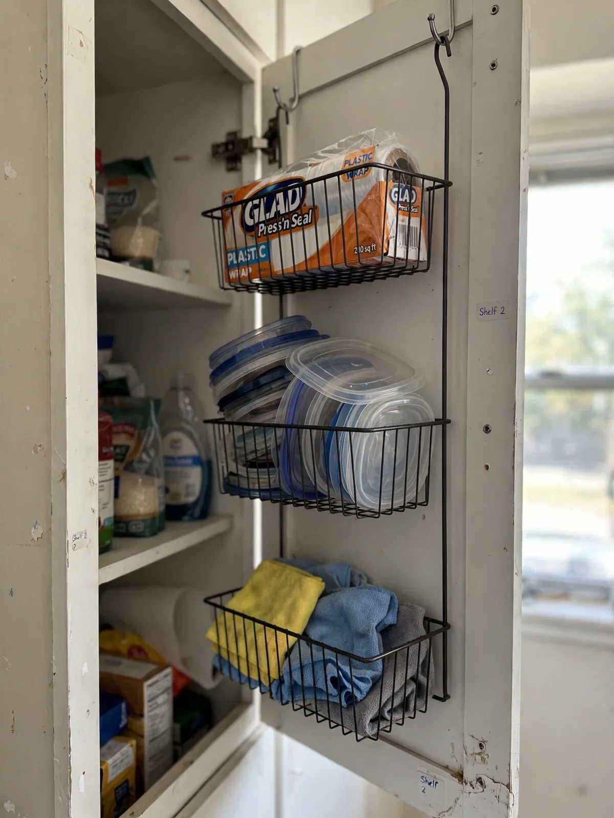 Wire baskets hanging over the inside of open kitchen cabinet doors in a rental apartment, holding wrap boxes lids and cleaning cloths on padded hooks