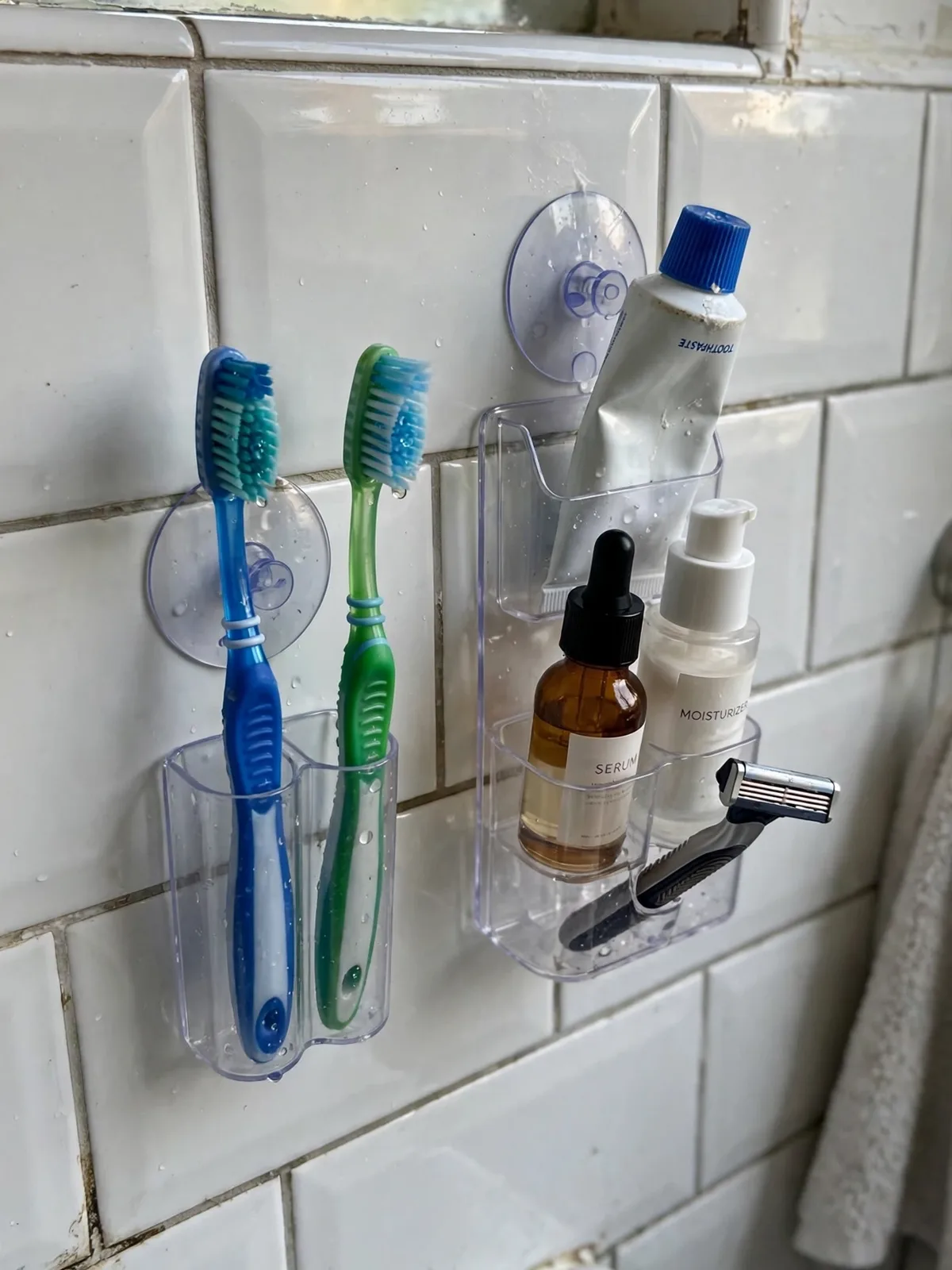 Clear suction-cup organizers stuck on glossy bathroom tiles in a small rental bathroom, holding toothbrushes toothpaste skincare bottles and a razor beside the sink mirror