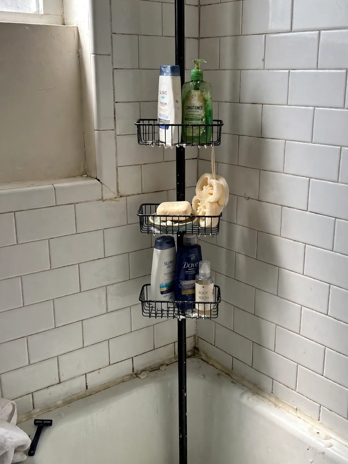 Tall tension-pole shower caddy standing floor to ceiling in the corner of a small rental bathtub, metal baskets holding shampoo bottles soap and a loofah neatly