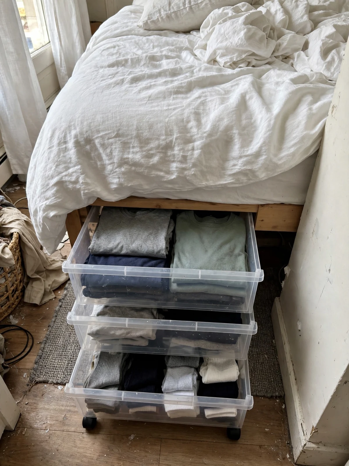 Stackable clear drawers under a bed pulled out slightly showing folded t-shirts and socks inside